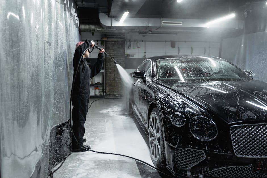 A person in a garage power washing a black car covered in soap and water spray.